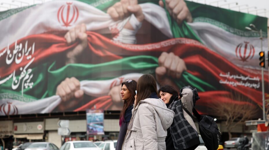 Young Iranian women walk past a large state propaganda poster in Tehran that reads "Iran is our homeland." January 2026. Photo: EPA/ABEDIN TAHERKENAREH